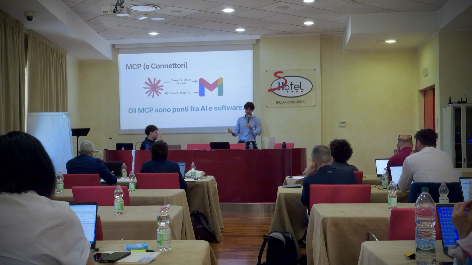 Riccardo Belli parla al microfono in una sala congressi, sullo schermo la slide "MCP (o Connettori): gli MCP sono ponti fra AI e software".